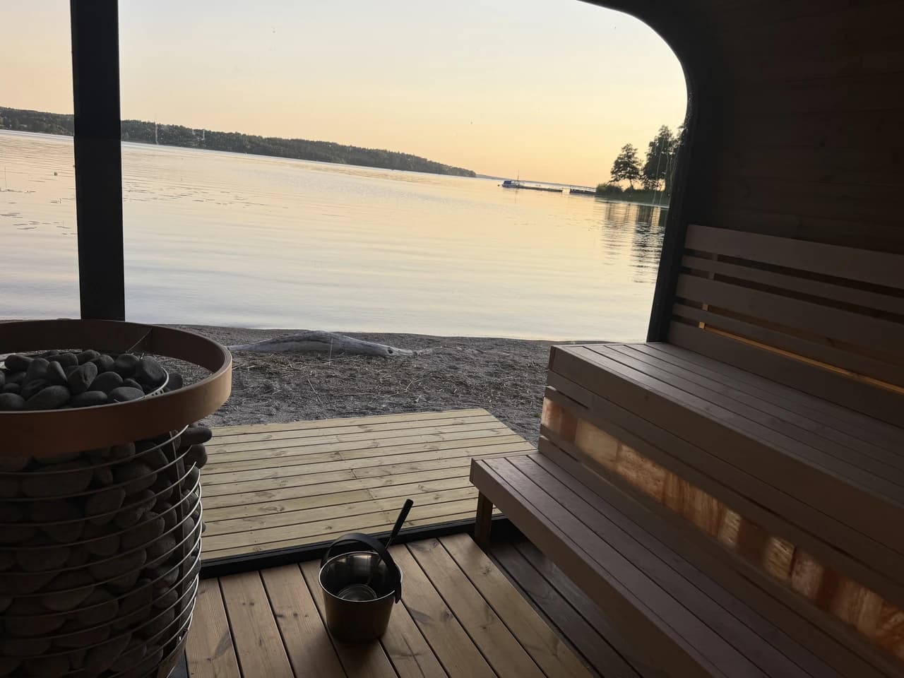 Utsikt från insidan av bastun på Kaggeholm i solnedgången, med stenfyllt aggregat, skopa och Mälaren utanför den öppna glasfronten