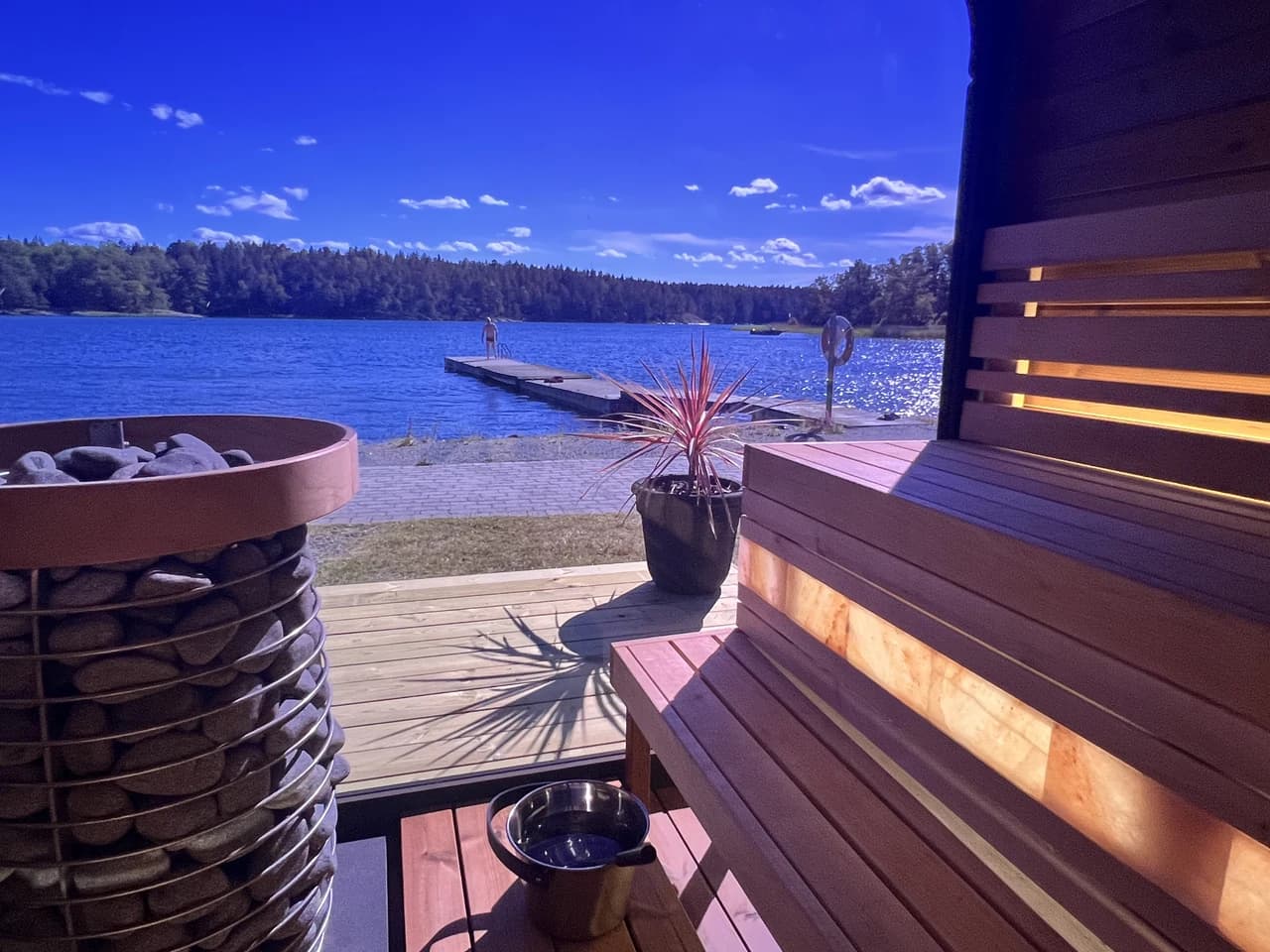 Utsikt från insidan av bastun vid Skåvsjöholm en solig sommardag, med stenfyllt aggregat, saltstensbelysta lavar och bryggan i skärgårdsvattnet