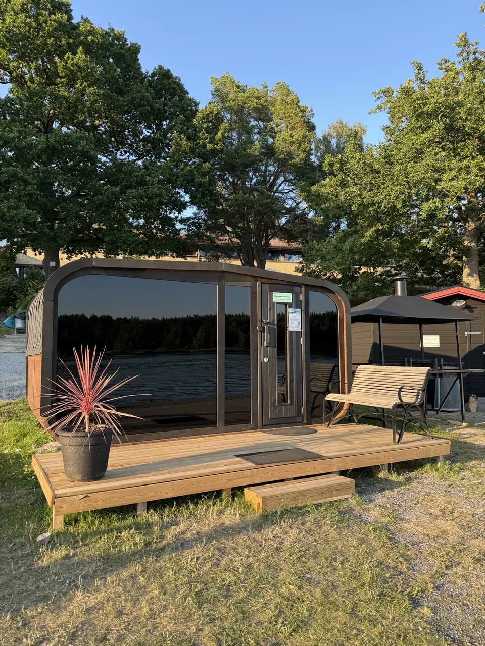 Skärgårdsbastun vid Skåvsjöholm på sitt trädäck i kvällssol, med stor glasfront, bänk och ekarna bakom