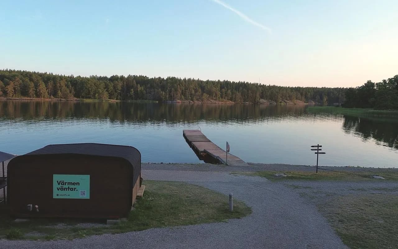 Skärgårdsbastu Åkersberga vid vattnet i kvällsljus, med bryggan och skogen på andra sidan viken