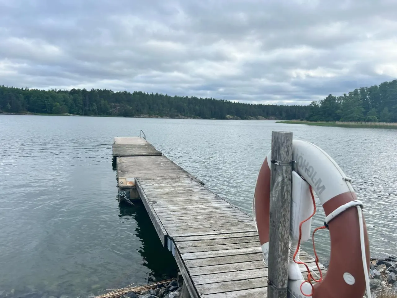 Badbryggan vid Skåvsjöholm ut i skärgårdsvattnet, med badstege längst ut, livboj på stolpen och den skogklädda stranden på andra sidan viken